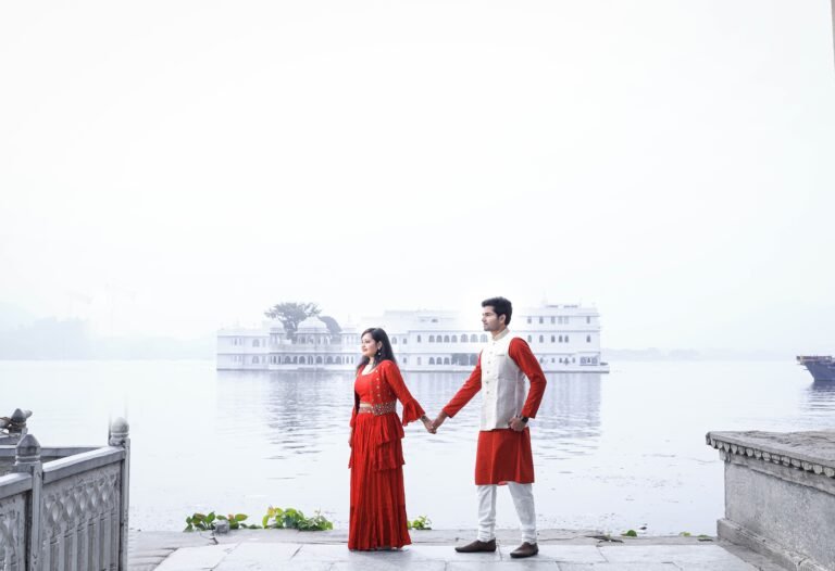Couple in red traditional outfits holding hands on a lake ghat with a white palace across the misty water