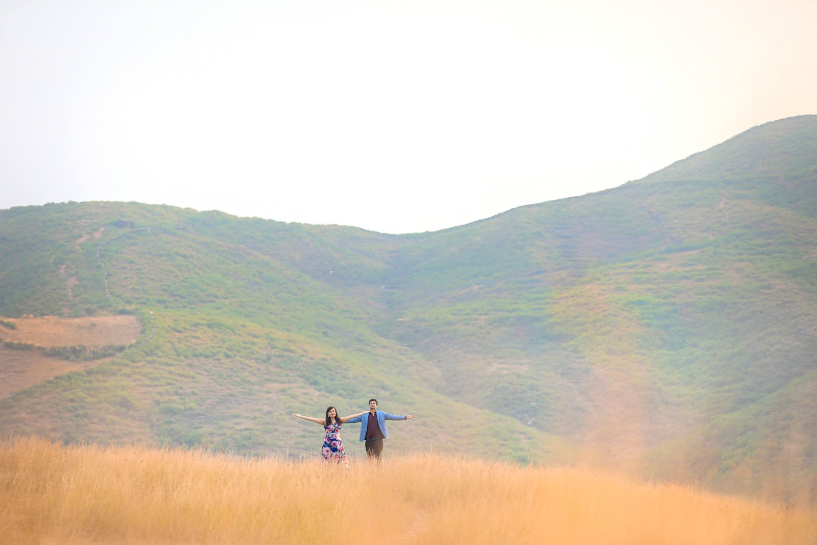 Couple standing with arms open in a wide golden field against green hills