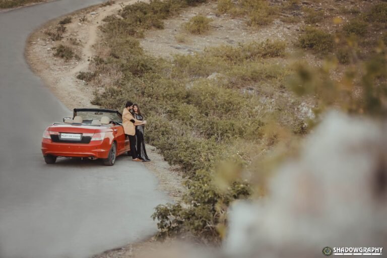 Couple standing by a red convertible on a winding hillside road during a pre-wedding shoot