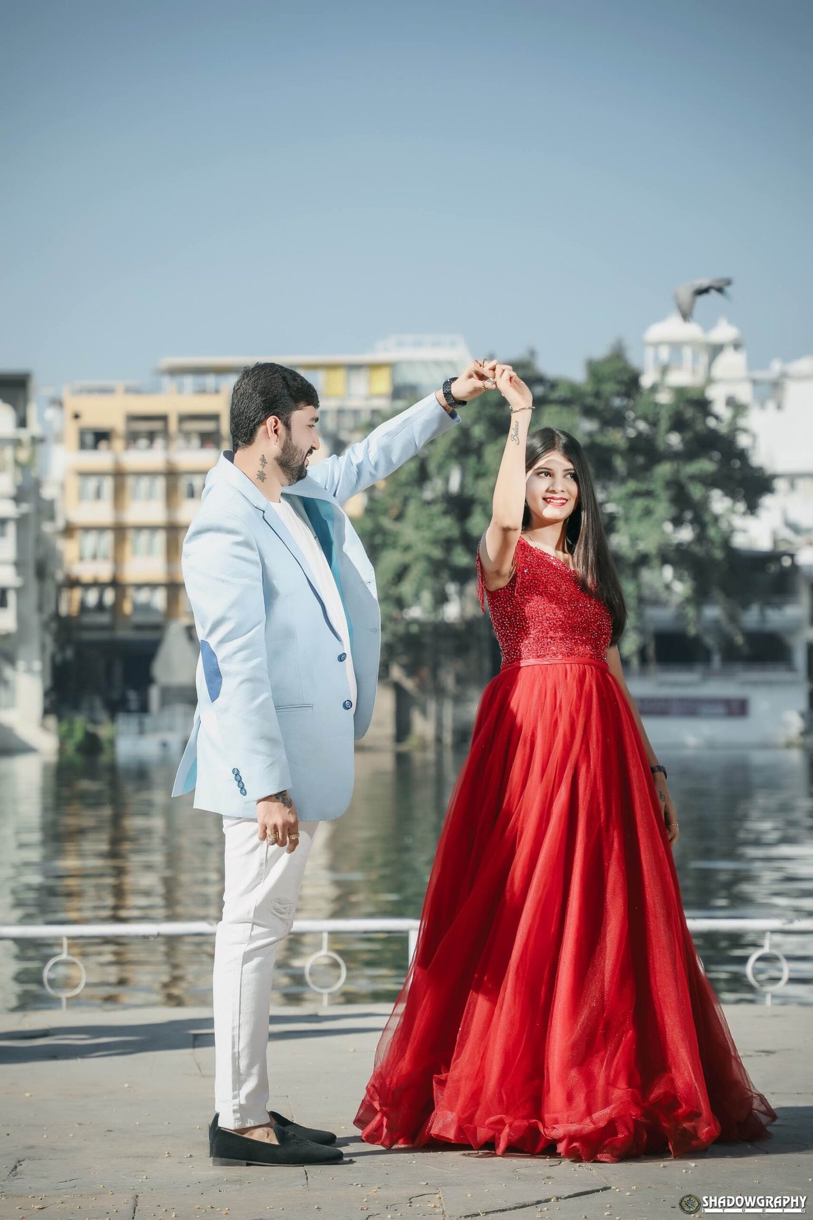 Couple in a red gown and light blue suit twirling on a lakeside promenade