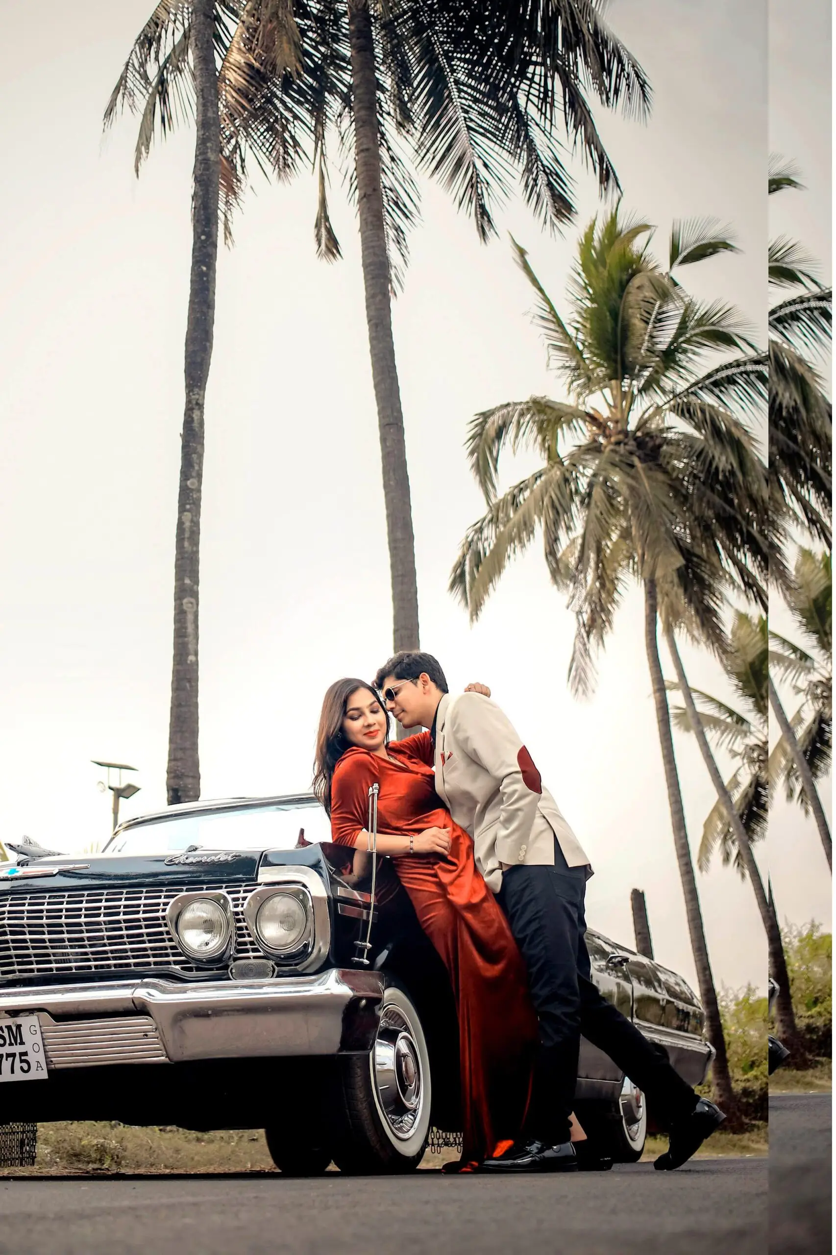 Couple in red and cream outfits leaning on a black vintage convertible under palm trees