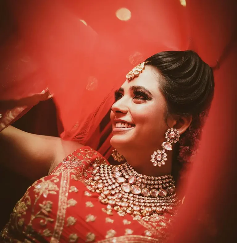 Bride in red lehenga with kundan choker, earrings and maang tikka framed by a red veil