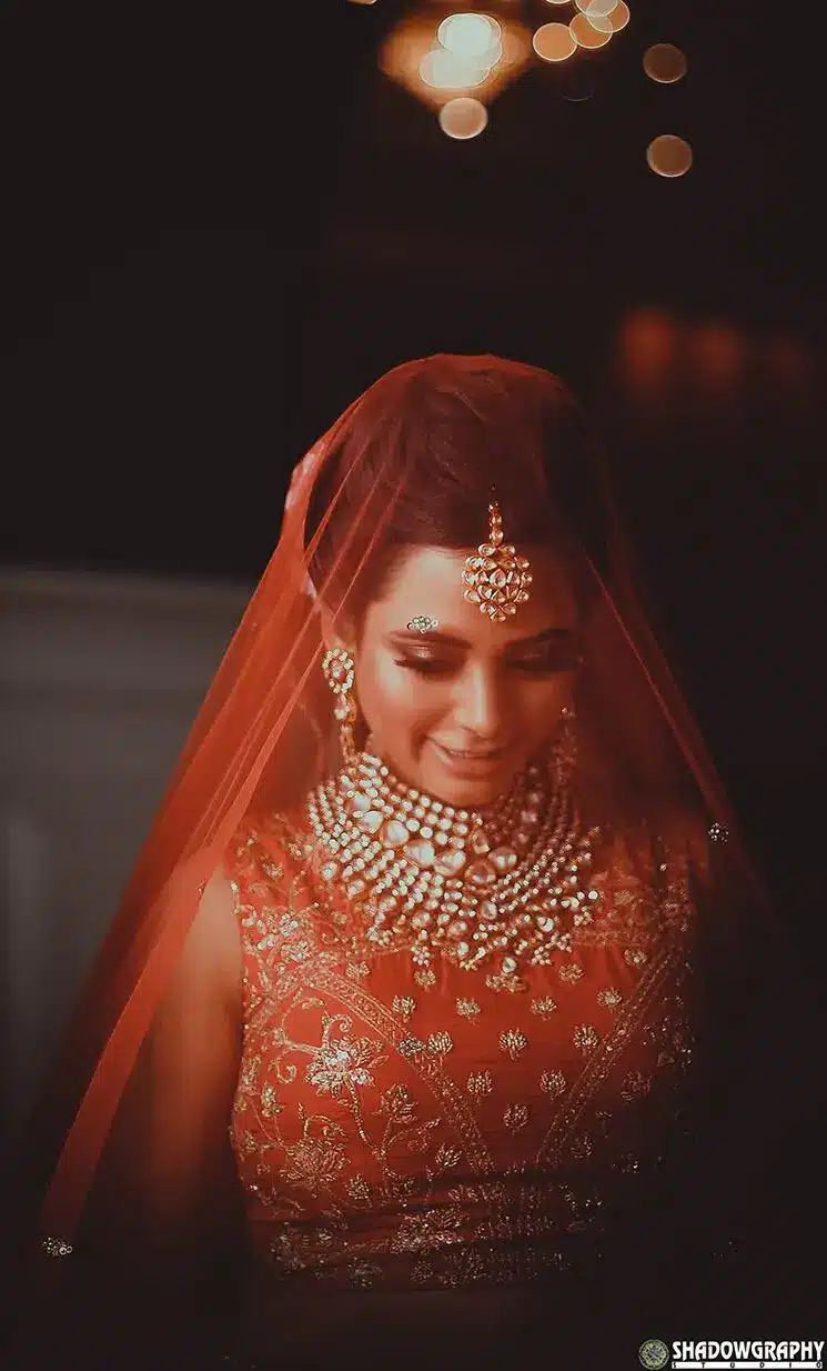 Bride in a red veil and jewellery photographed indoors under warm light with soft bokeh behind her
