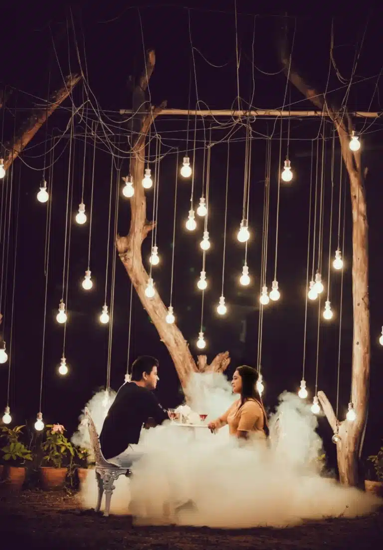 Couple seated under hanging Edison bulbs with soft fog on a night set, pre-wedding shoot near Indore, by Shadowgraphy Studio