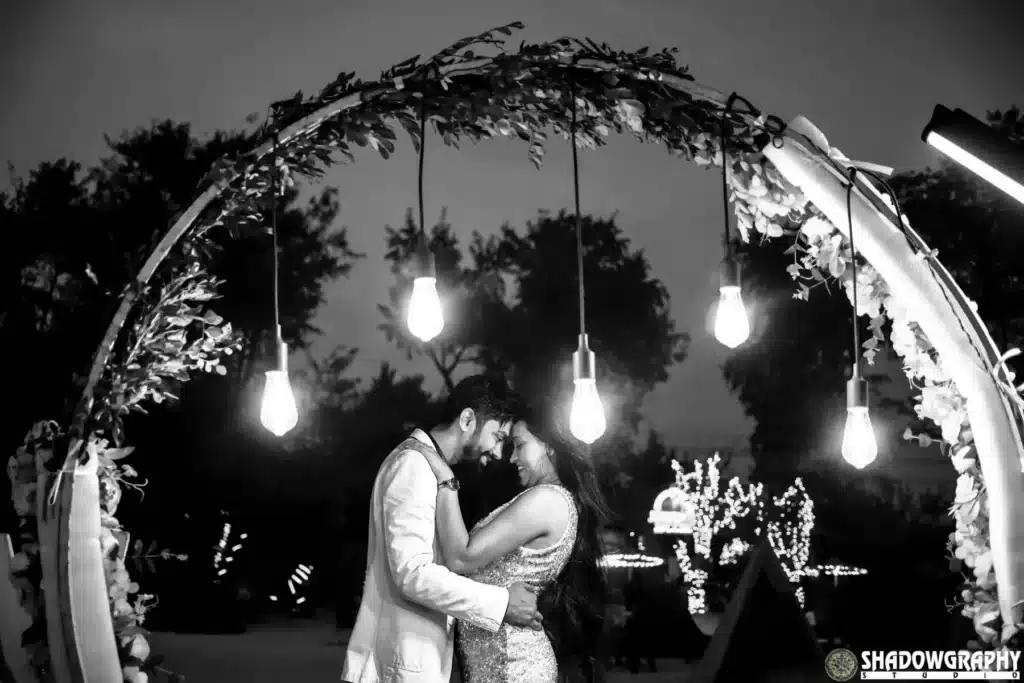 Black and white pre-wedding photo of a couple embracing under a floral arch with hanging bulbs