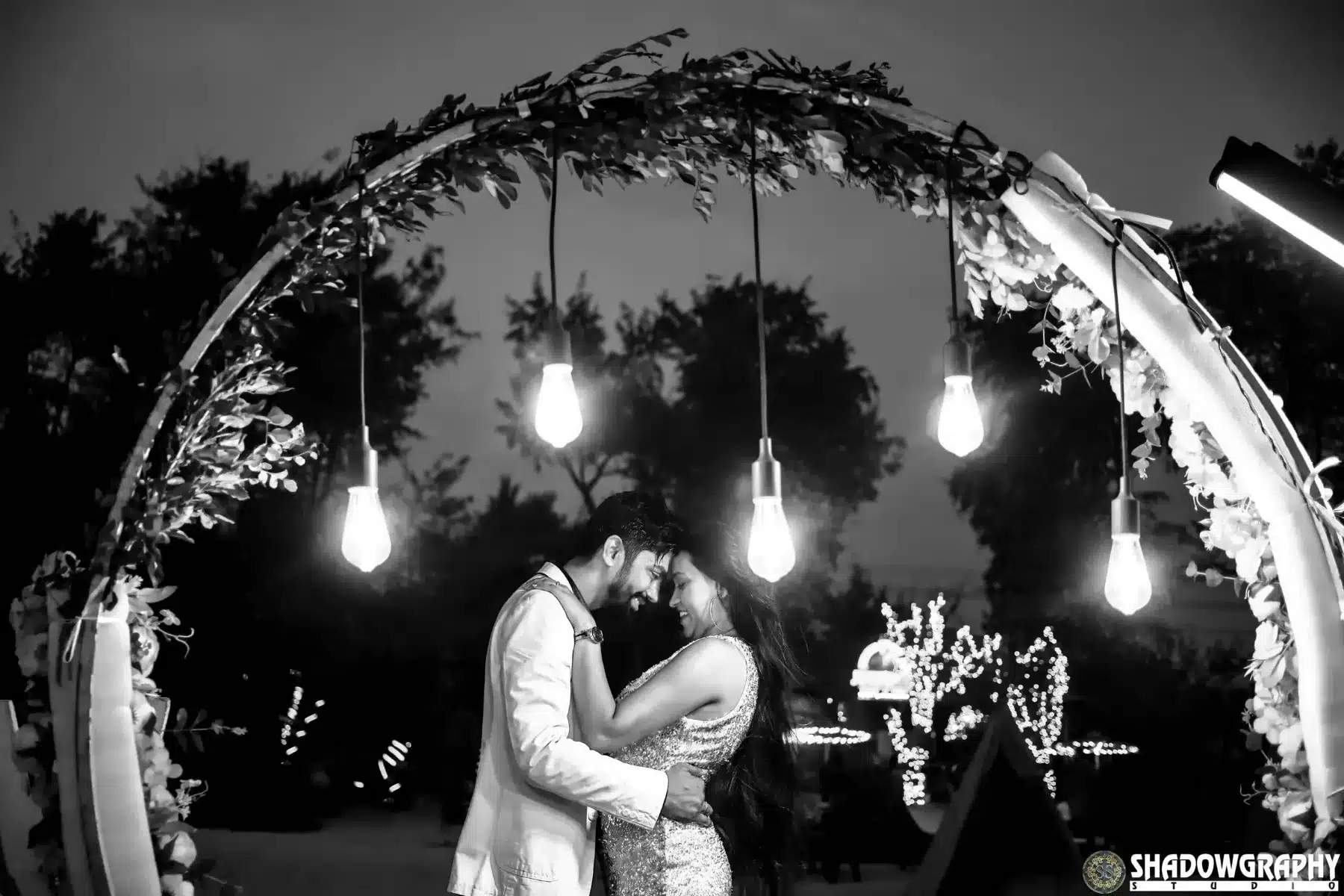 Black and white pre-wedding photo of a couple embracing under a floral arch with hanging bulbs