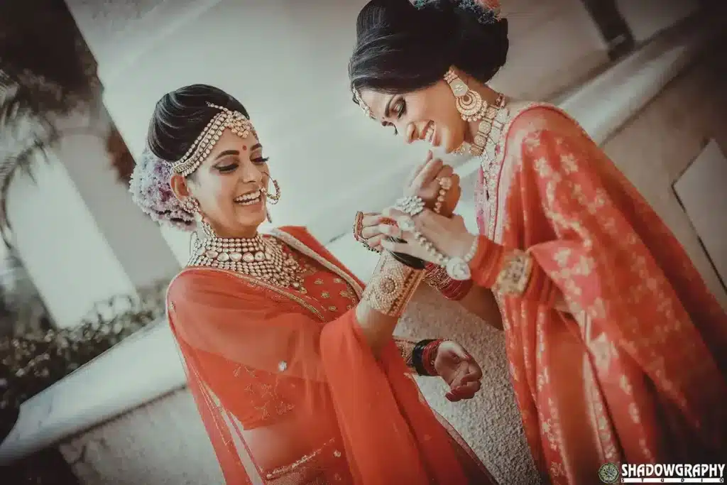 Two women in orange bridal outfits sharing a candid moment during bridal preparation