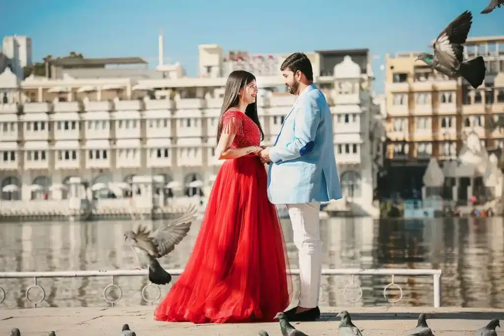 Pre-wedding couple in a red gown and blue kurta posing on a lakeside promenade with a palace backdrop