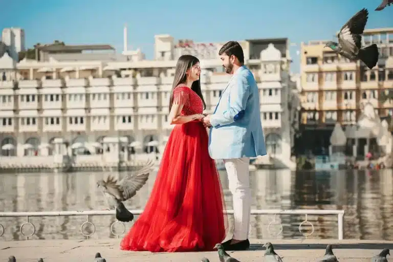 Pre-wedding couple in a red gown and blue kurta posing on a lakeside promenade with a palace backdrop
