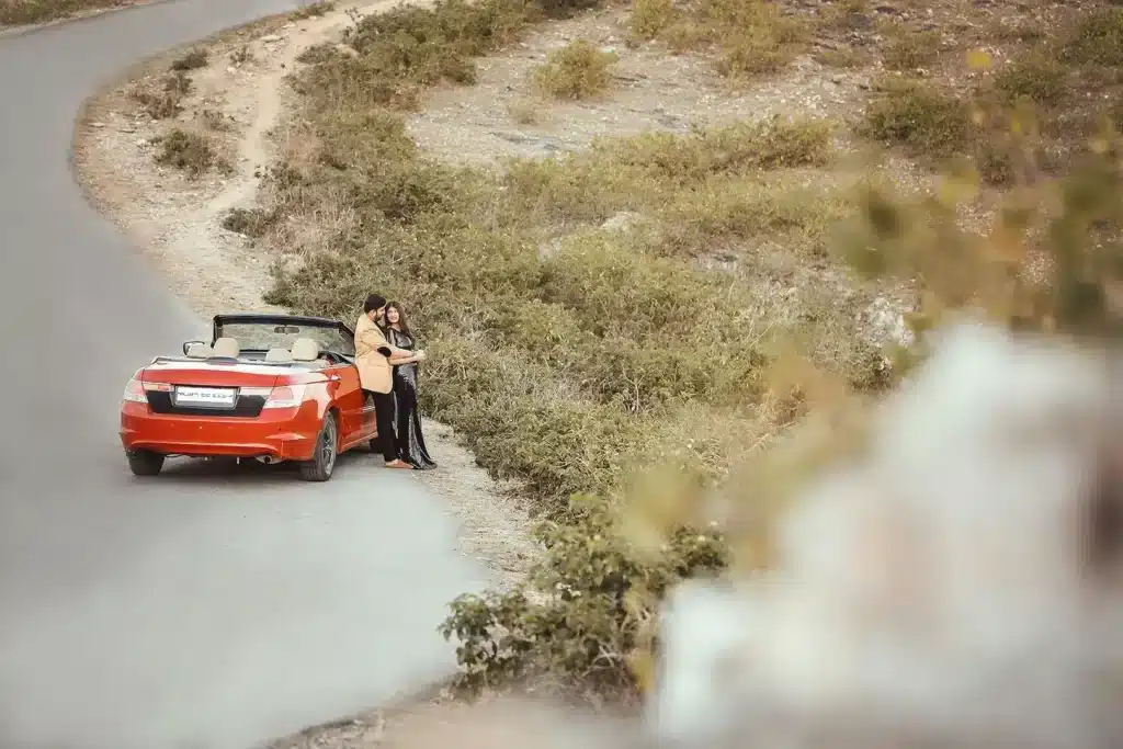 Pre-wedding couple posing beside an orange convertible car on a hillside road