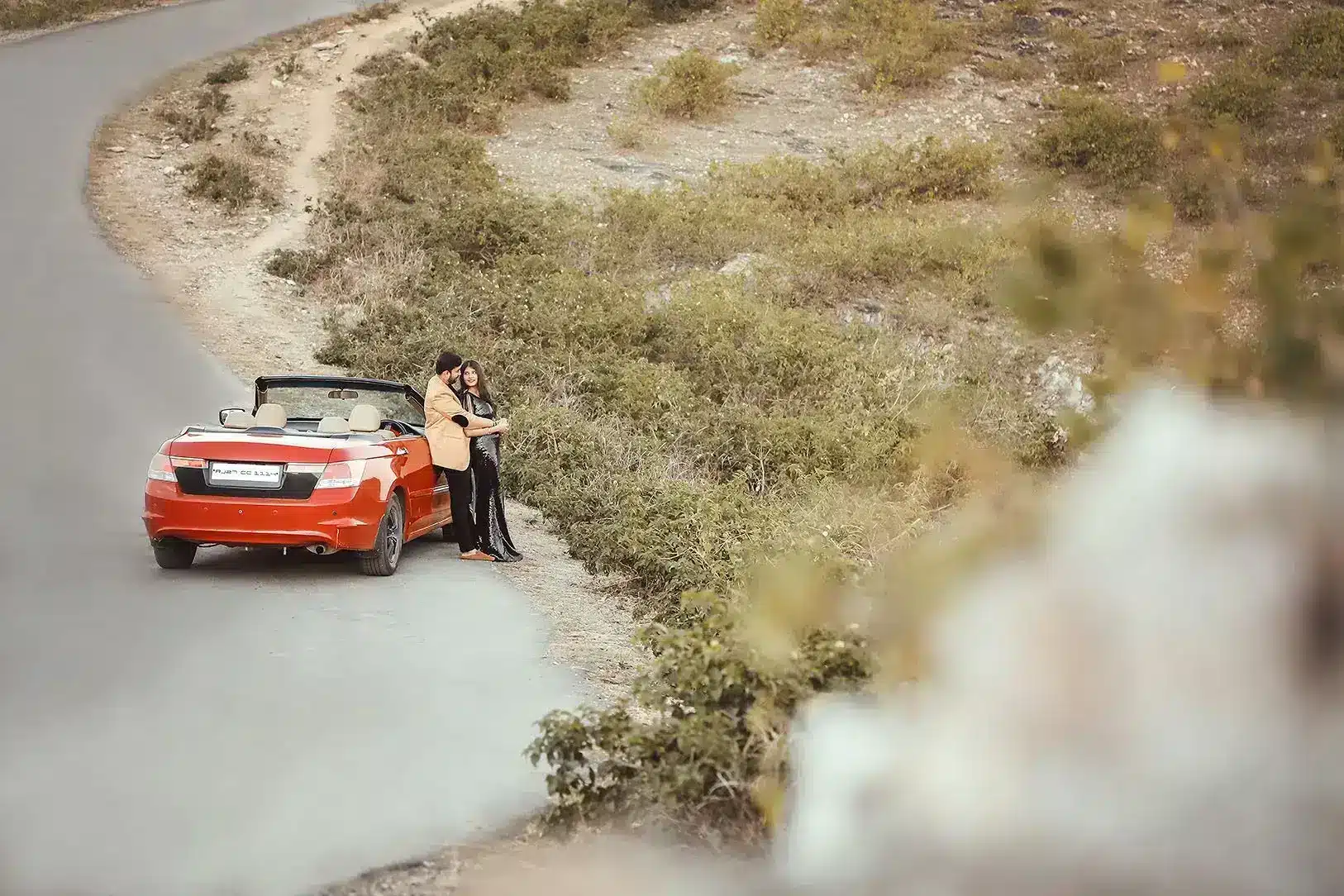 Pre-wedding couple posing beside an orange convertible car on a hillside road