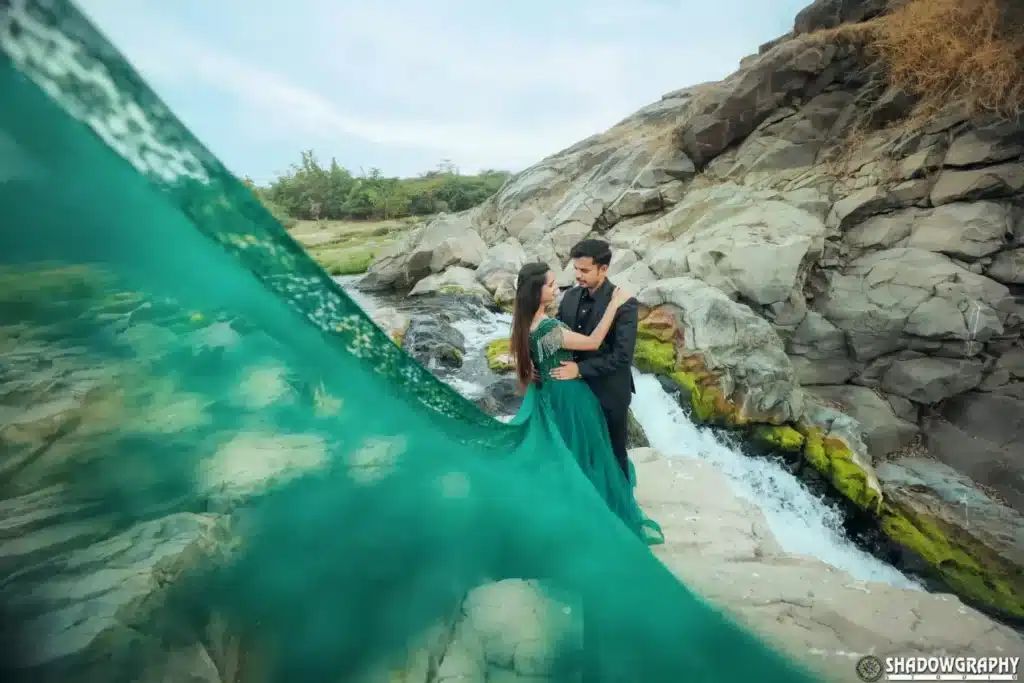 Pre-wedding couple standing on rocks with a long teal gown flowing over the water